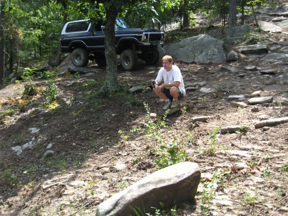 Man crouching near a 1978 Ford Bronco on a rocky, wooded trail