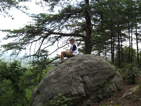 1978 Ford Bronco - A person sits on a large boulder in a wooded area, looking out over a distant landscape.