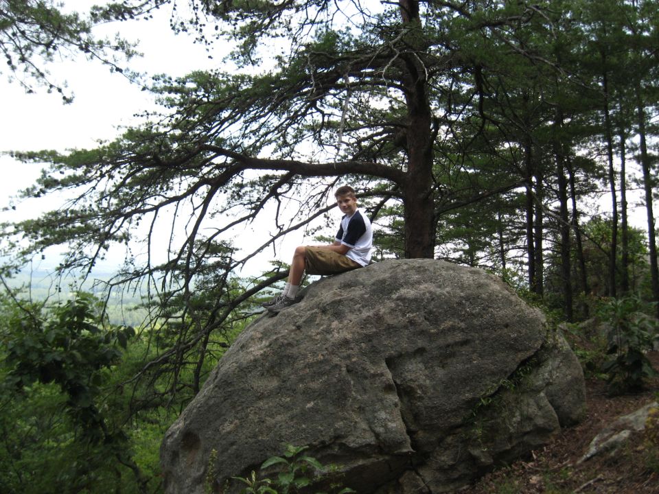 Boy sits on a large boulder under a pine tree, with a forest and distant valley in the background