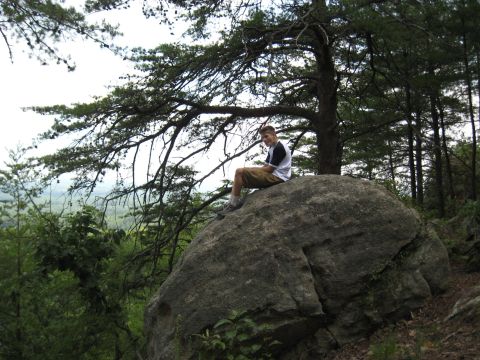 1978 Ford Bronco - A person rests on a large rock formation amidst trees, with a view of the distant landscape.