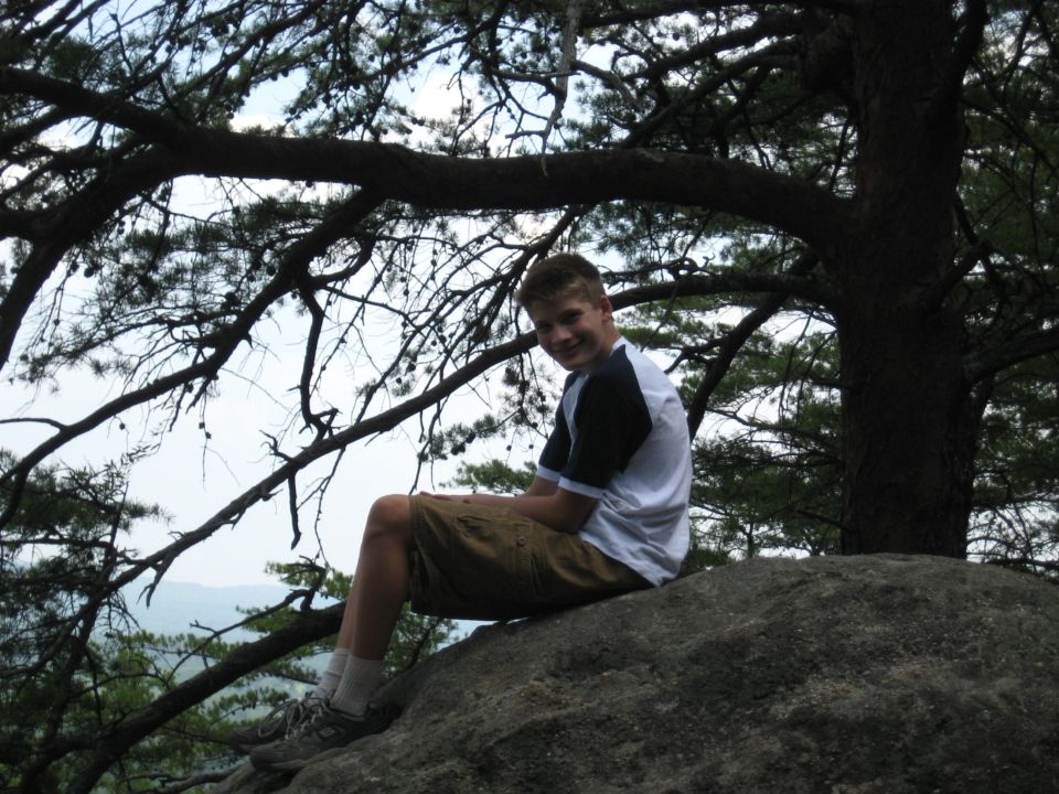 Young man in a t-shirt and shorts sits on a large rock under a tree, smiling at the camera