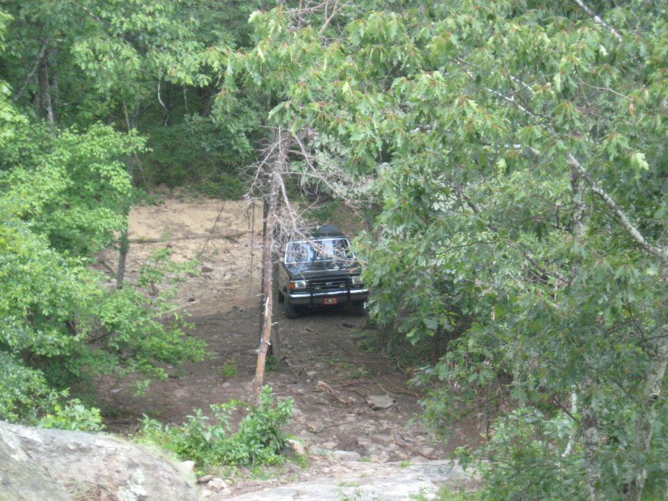 Black SUV with a red license plate driving on a muddy, rocky path surrounded by trees