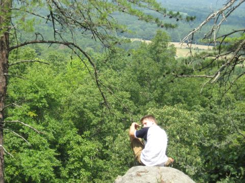 1978 Ford Bronco - A person contemplates the view from a rocky outcrop amidst dense trees.