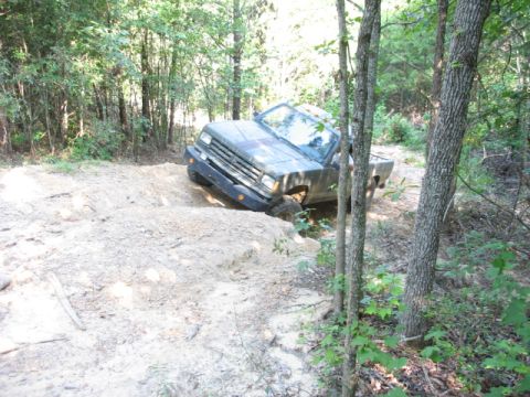 1978 Ford Bronco - A dark-colored truck navigates a challenging dirt path through a wooded area, with one wheel significantly higher than the others.