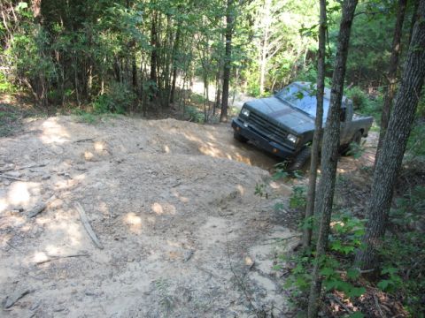 1978 Ford Bronco - Pickup truck driving off-road through a wooded trail with a sandy incline.