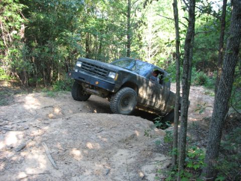 1978 Ford Bronco - A dark pickup truck drives off-road on a dirt path surrounded by trees.