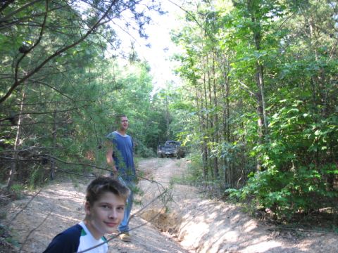 1978 Ford Bronco - A truck drives down a dirt road in a wooded area, with two people nearby.