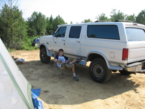 1978 Ford Bronco - A boy sits in a chair next to a white Ford Bronco SUV parked in a dirt lot with trees in the background.
