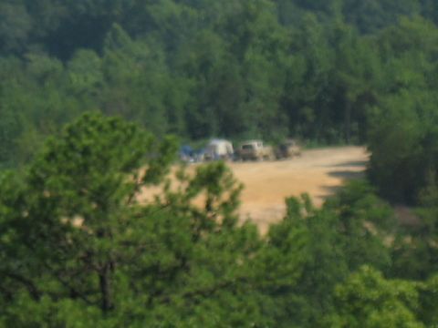 1978 Ford Bronco - A collection of vehicles and camping tents are visible in a clearing amidst a dense forest.
