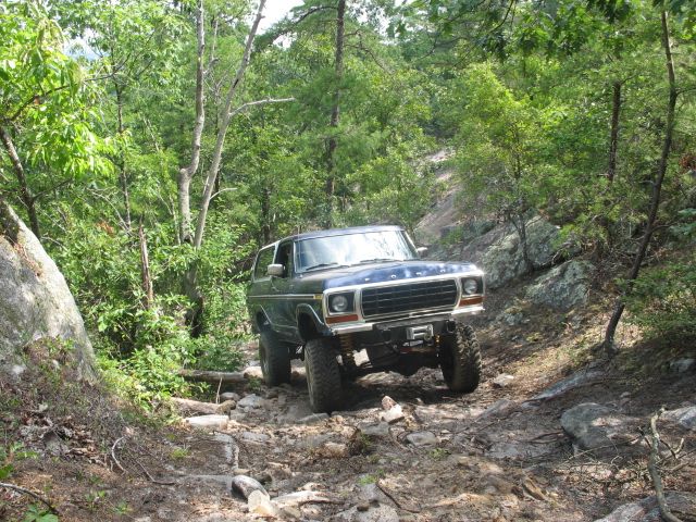 Blue 1978 Ford Bronco navigating a steep, rocky off-road trail with trees on either side