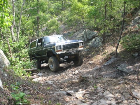 1978 Ford Bronco - A lifted 1978 Ford Bronco navigates a challenging off-road path through a dense forest.