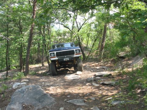 1978 Ford Bronco - A blue 1978 Ford Bronco navigates a challenging, rocky off-road trail through a wooded area.