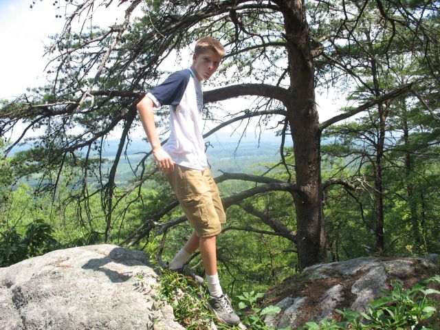 Young man in a t-shirt and cargo shorts stands on a rock ledge with trees and a valley in the background