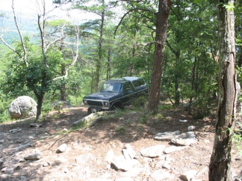 1978 Ford Bronco - A 1978 Ford Bronco is parked on a trail surrounded by trees and rocks, with a distant view of hills.