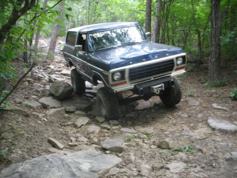 1978 Ford Bronco - A blue 1978 Ford Bronco navigates a challenging, rocky off-road path surrounded by trees.