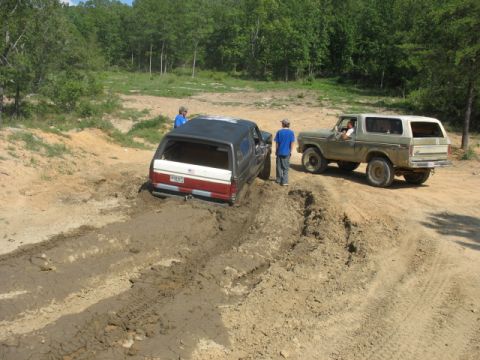 1978 Ford Bronco - Two SUVs are stuck in the mud on a dirt track, with two people observing.