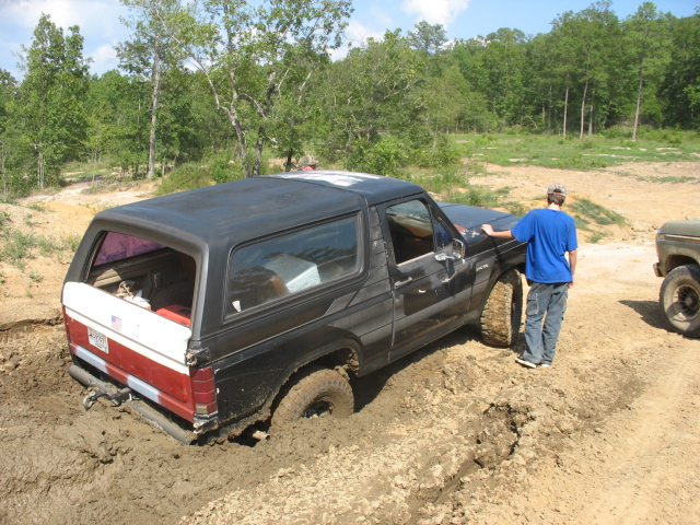Black Ford Bronco stuck in mud with a person standing next to it. Another vehicle is visible to the right