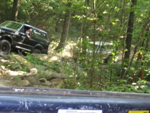 1978 Ford Bronco - A black SUV tackles a challenging rocky path in a wooded area, with another vehicle partially visible behind it.