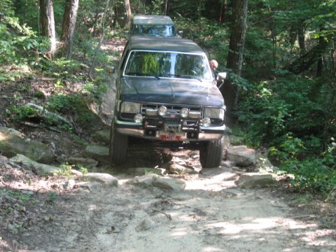 1978 Ford Bronco - A dark blue Ford Bronco drives over a rocky path in a wooded area, with another vehicle visible behind it.