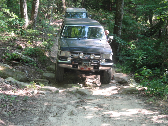 Dark blue Ford Bronco navigating a rocky trail, front view. Another vehicle follows behind