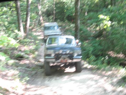 1978 Ford Bronco - Two off-road vehicles navigate a wooded trail, with the lead vehicle a winch and auxiliary lights.