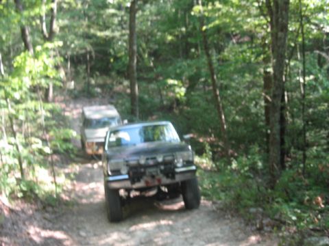 1978 Ford Bronco - Two SUVs navigate a dirt path through a forest. The lead vehicle is equipped for off-road use.