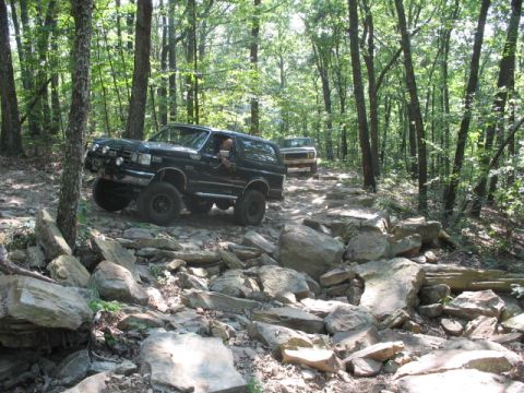 1978 Ford Bronco - Two SUVs drive through a challenging, rocky path surrounded by dense trees.