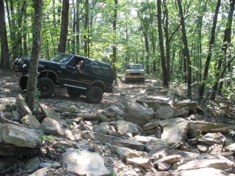 1978 Ford Bronco - Two off-road vehicles traverse a challenging, boulder-strewn path through a dense forest.