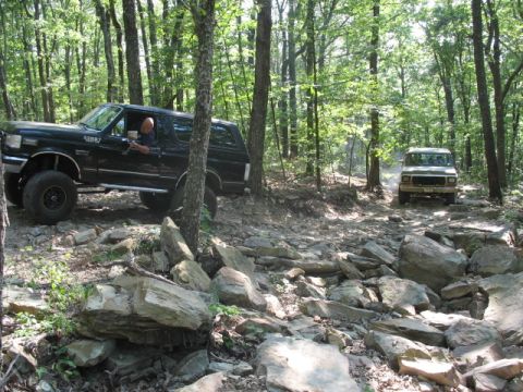 1978 Ford Bronco - A black Ford Bronco drives through a challenging, rocky path in a wooded area, with a second vehicle visible further down the trail.