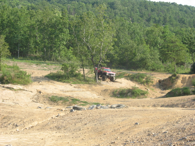Red off-road SUV on dirt track, front 3/4 view, trees and hills in background