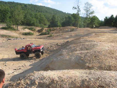 1978 Ford Bronco - Custom red off-road truck navigates a challenging dirt terrain with a wooded hill in the background.