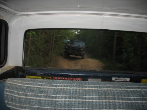 1978 Ford Bronco - A black SUV is seen driving on a dirt road surrounded by trees, viewed from inside another vehicle.