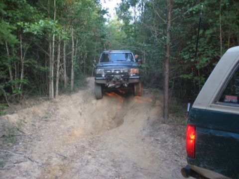 1978 Ford Bronco - Two SUVs navigate a challenging dirt trail through a wooded area, with one vehicle descending into a rut.