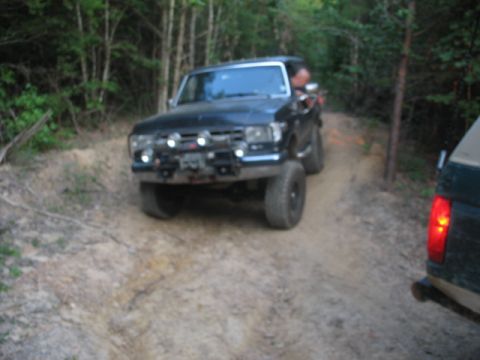 1978 Ford Bronco - A black SUV drives on a dirt path surrounded by trees, with another vehicle visible to the right.