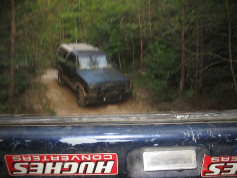 1978 Ford Bronco - A blue SUV with a camper shell navigates a muddy dirt road through a wooded area, with a Hughes Converters sticker visible on the vehicle i…