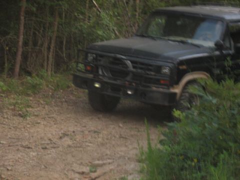 1978 Ford Bronco - A black SUV with a brush guard drives on a dirt road surrounded by trees and foliage.