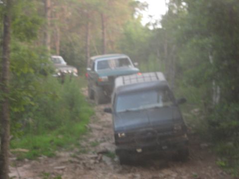 1978 Ford Bronco - Several SUVs navigate a challenging off-road path through a dense forest.