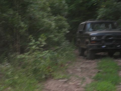 1978 Ford Bronco - A dark SUV drives on a dirt path surrounded by trees, captured with motion blur.
