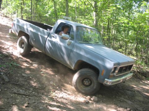 1978 Ford Bronco - A person drives a light blue pickup truck up a steep, rocky dirt path surrounded by trees.