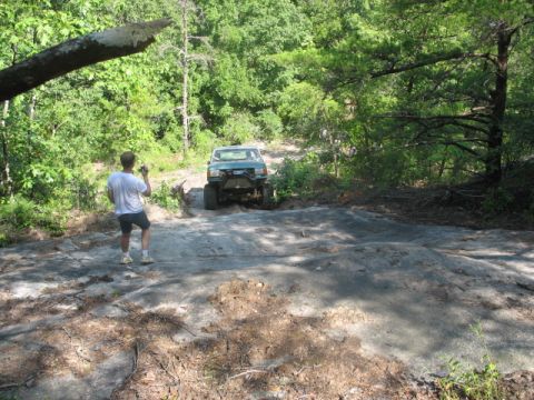 1978 Ford Bronco - A person films an off-road vehicle navigating a challenging rocky incline in a wooded area.