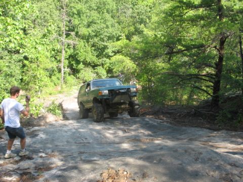 1978 Ford Bronco - A person photographs a green SUV navigating a challenging rocky terrain in a wooded area.