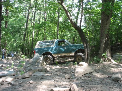 1978 Ford Bronco - A teal Ford Bronco is shown navigating a challenging, rocky trail in a wooded area.