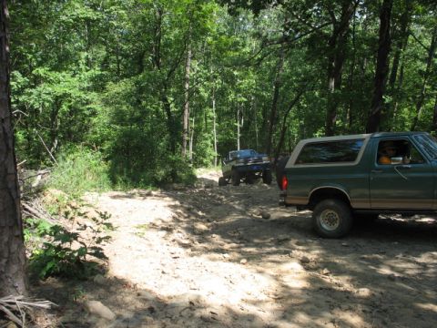 1978 Ford Bronco - A green SUV navigates a rocky, wooded trail with another vehicle ahead.
