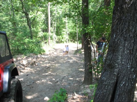 1978 Ford Bronco - People walk on a dirt trail in a wooded area, with a red vehicle partially visible on the left.