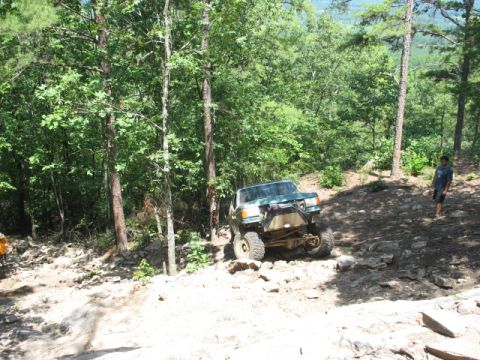 1978 Ford Bronco - An off-road SUV navigates a steep, rocky trail surrounded by dense forest.