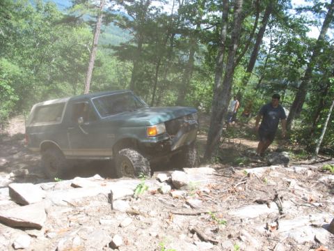 1978 Ford Bronco - A green SUV is shown off-roading on a rocky trail surrounded by trees.