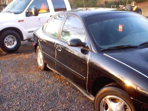 1999 Ford Taurus - A black sedan is parked next to a white truck on a gravel surface.