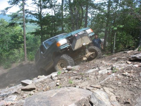 1978 Ford Bronco - A modified green SUV navigates a steep, rocky incline in a wooded area.