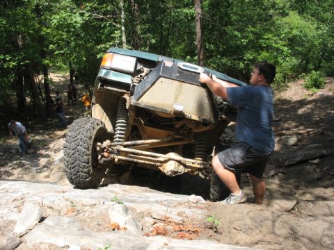 1978 Ford Bronco - A person helps push a green off-road SUV navigating a difficult rocky trail with other vehicles and people in the background.