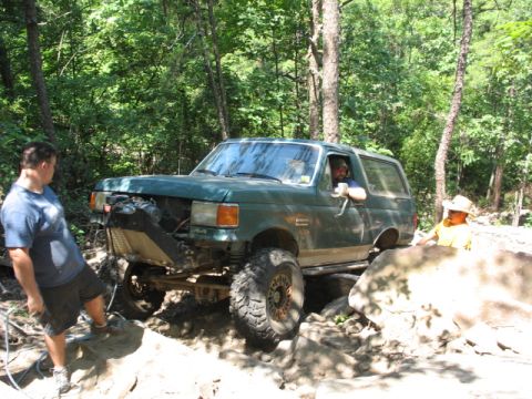 1978 Ford Bronco - A green Ford Bronco is shown off-roading on a rocky trail with trees in the background.